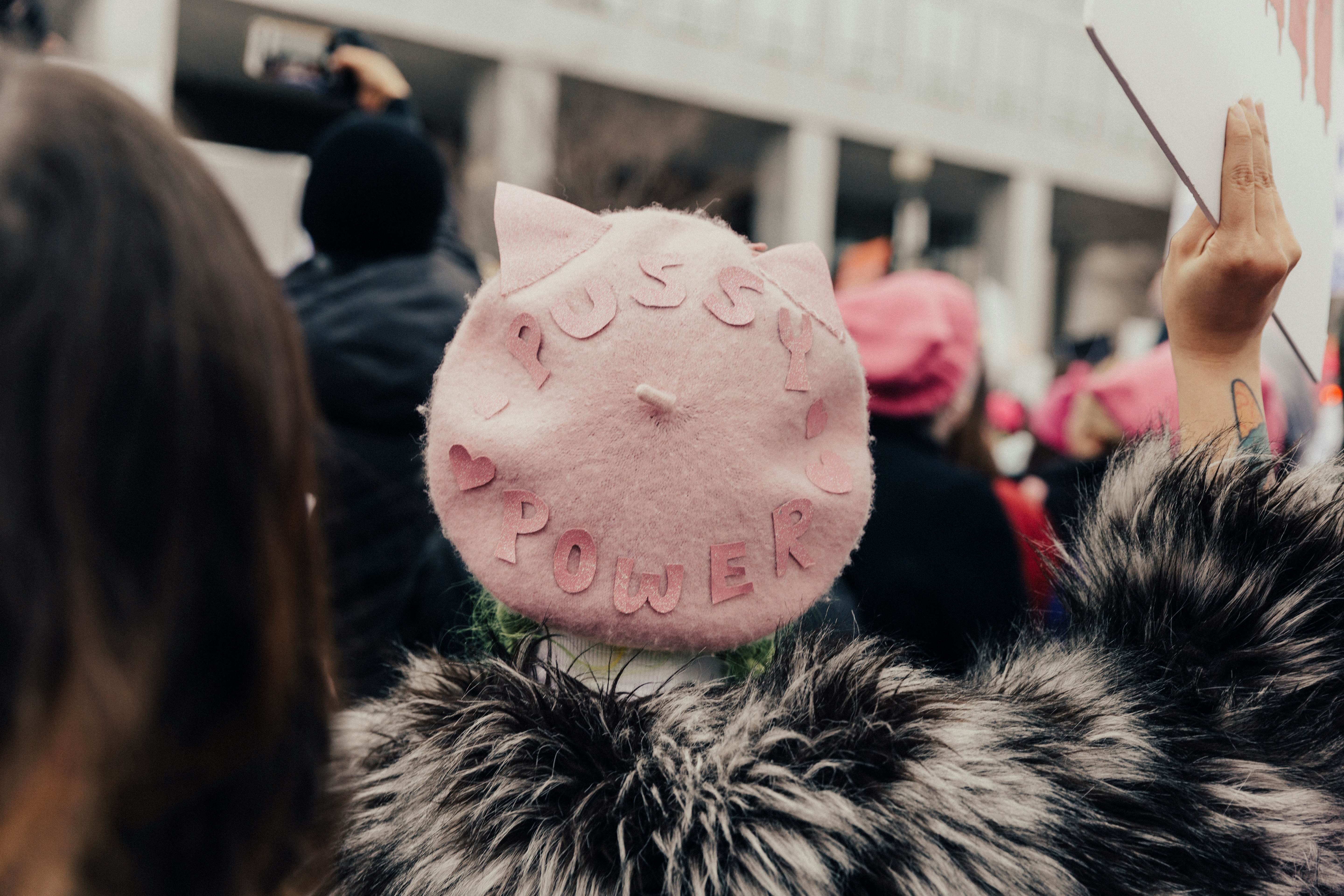 selective focus photography of person wearing pink fleece Pussy Power cap raising right hand